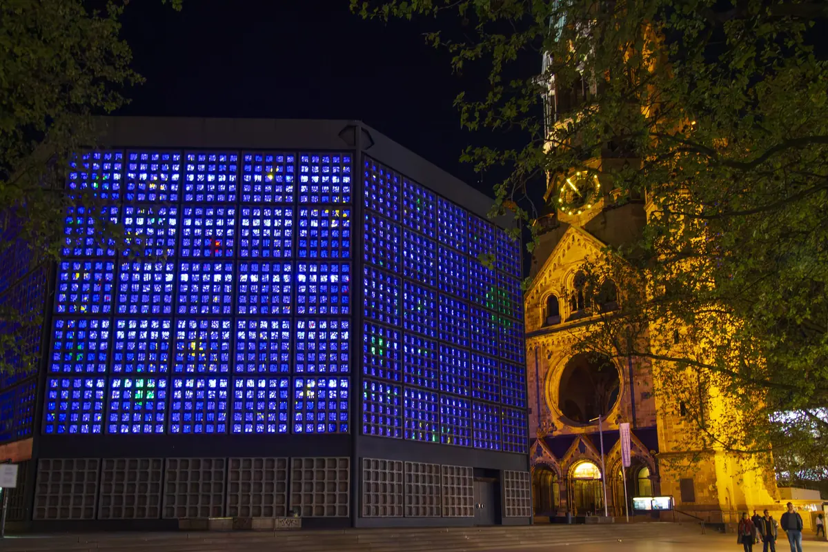 The new Memorial Church at night, its blue glass walls glowing beside the floodlit sandstone tower of the old church and a lit street in front